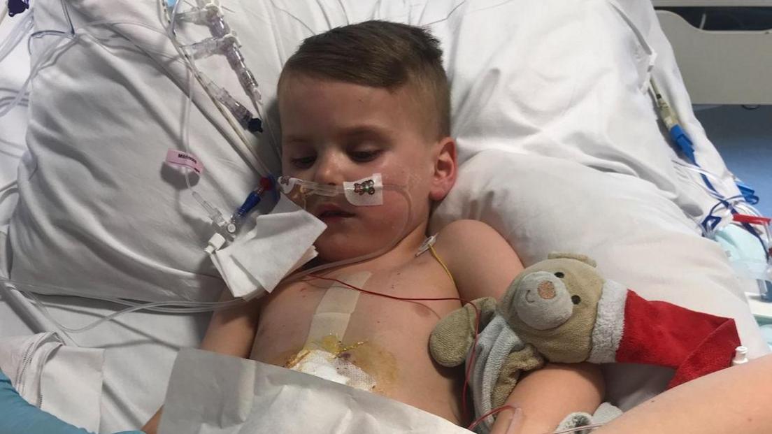 Jayden lying in a hospital bed with arm around a teddy. He has oxygen tubes attached to his nose and a bandage is visible on his stomach. 