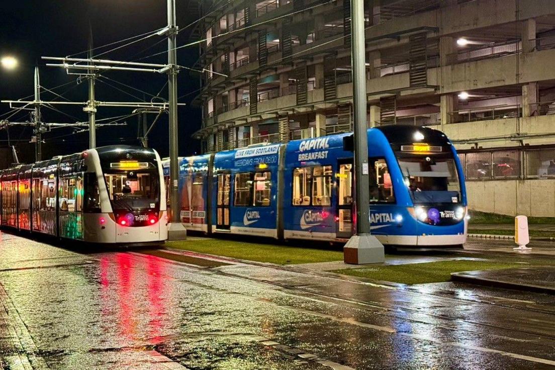 Two trams one white, the other blue, are parked in a street. The ground is wet and they are illuminated in the dark by street lights.