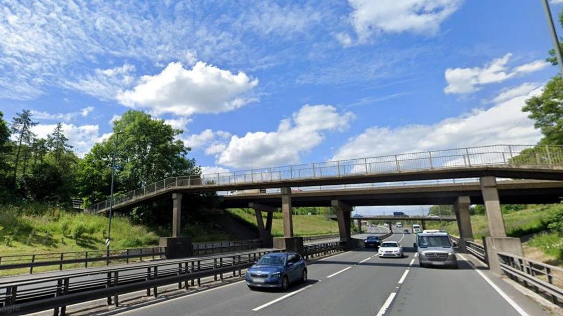 A Google Maps photograph of the A19 at Norton Interchange. A blue car is travelling in the left lane, a white car in the middle one and a grey van is in the right lane. There is a footbridge over the road and other vehicles in the distance, including a lorry. There are trees on the side of the road and the weather is sunny. 