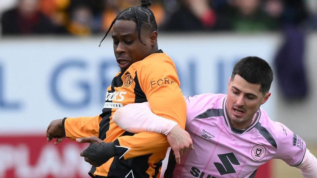 Pelly Ruddock Mpanzu links arms with Jon Mellish of MK Dons as they challenge for the ball