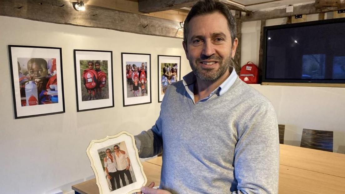 Luke Simon stood in his house holding a photograph of him and his brother Piers when they were younger. There are photos of children on the wall who have been helped through his charity.