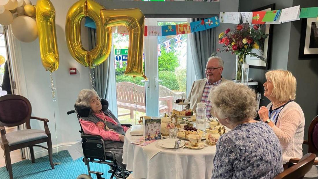 Ms Jones sitting at a table where a high tea has been set. There are three people sitting with her and in the background there are balloons and lots of cards hanging on a string