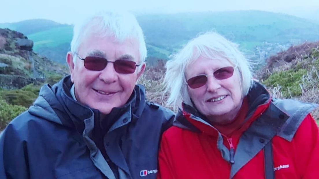 Head and shoulders photo of Eric and Val in happier times, both smiling to the camera. They both have grey hair and are wearing sunglasses and waterproof jackets. They are on a hill with more hills seen in the background. 