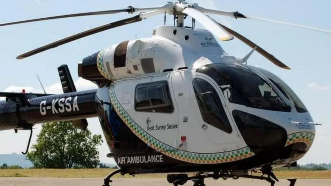 A black and white ambulance, with Kent Surrey Sussex Air Ambulance written on it. The ambulance is on the ground, with grass and a tree visible behind it