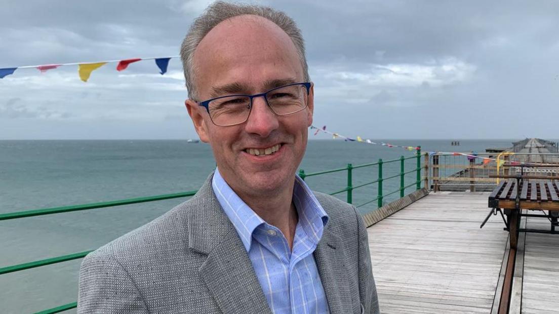 Tim Baker wearing glasses, a blue shirt and grey suit jacket, smiling while standing on the Queen's Pier in Ramsey on a cloudy day.