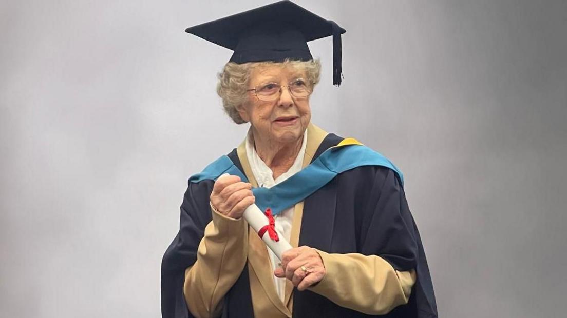 An elderly woman with curly grey hair holds a white scroll with both hands while wearing a navy blue gown and navy blue mortarboard on her head. 