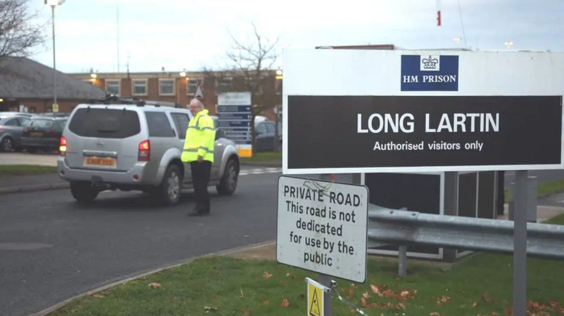 Image of the entrance to HMP Lartin which shows a large rectangular white and black sign which shows the name of the prison. A man in a yellow high-vis jacket is seen in the foreground of the image stood next to a silver SUV car.
