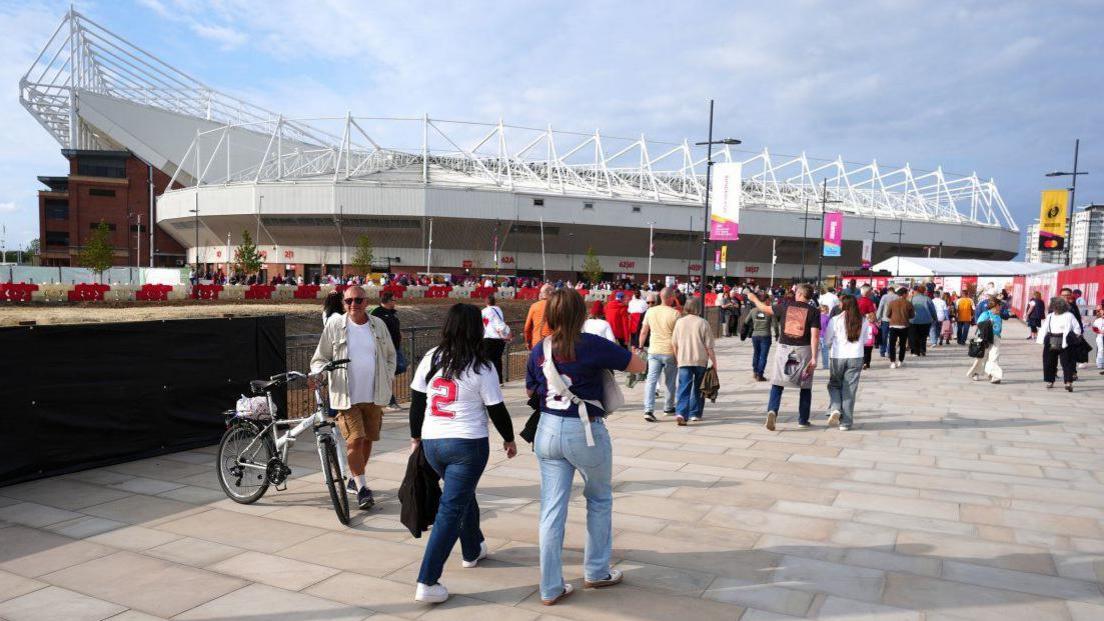 Rear view of a large number of people walking across a paved area towards a large arena building - Sunderland's Stadium of Light.