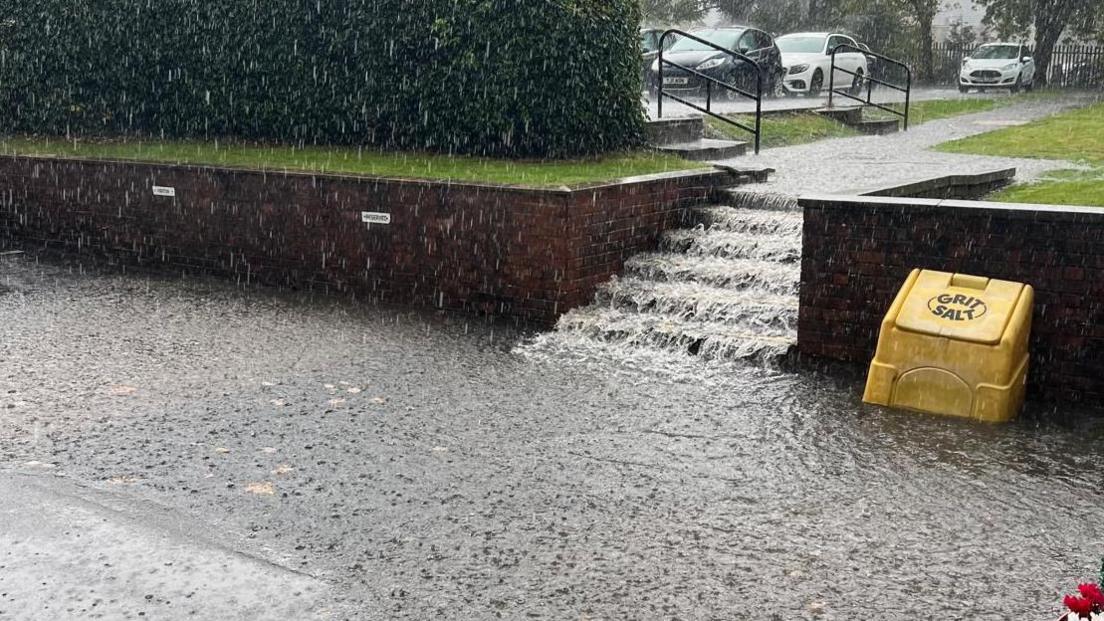 Water gushing down the steps of the school, from a grassed area - there is a gritter salt box on the right