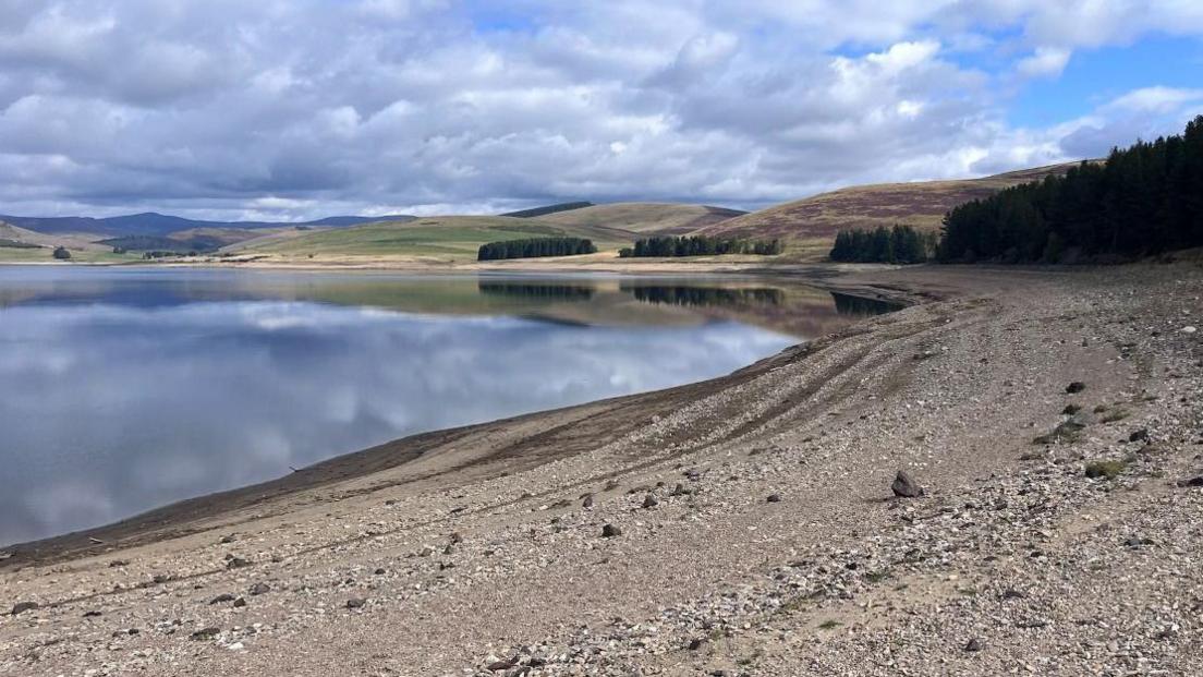 A reservoir with a stony beach in the foreground and hills in the background