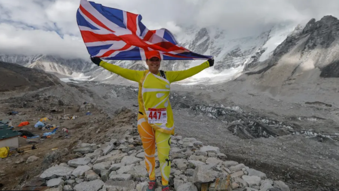 A Sally Orange wearing a yellow and orange citrus costume while hold the union Jack flag on Mount Everest.