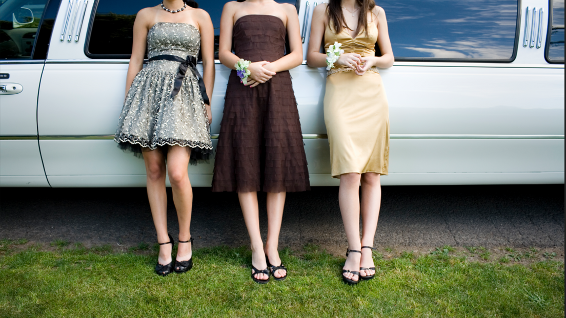 Three girls in Prom outfits and heels lean against a long car by some grass. You cannot see their faces.