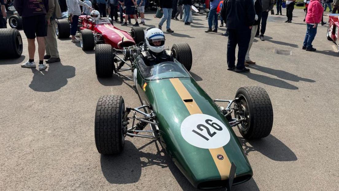 A single-seated green smaller racing Formula-1 style car with the number 126 on the front on a tarmac forecourt. A driver is sitting inside, wearing a dark jumpsuit and a white helmet. A similar red car can be seen behind it and the bodies and torsos of people are seen standing around the vehicles talking.