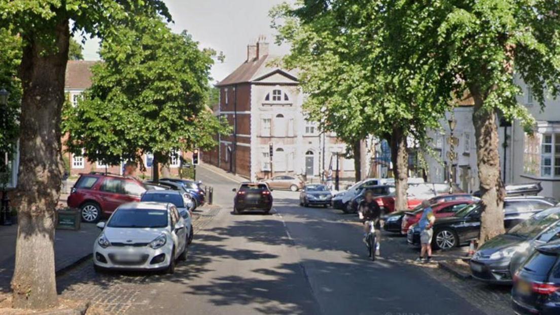 A street with cars and trees along both sides and a tall house in the background