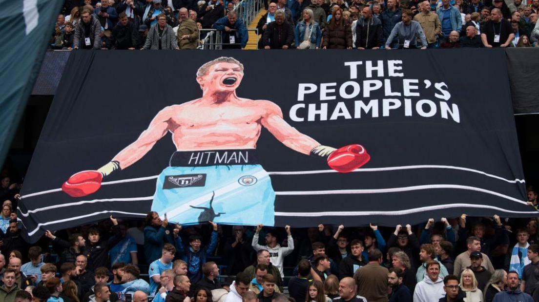 A large rectangular banner is held up by fans at a Manchester City game in honour of Ricky Hatton. Against the black background there is a depiction of a triumphant Hatton, wearing sky blue Manchester City shorts and red boxing gloves. White writing in large capital letters declares him to be The People's Champion.