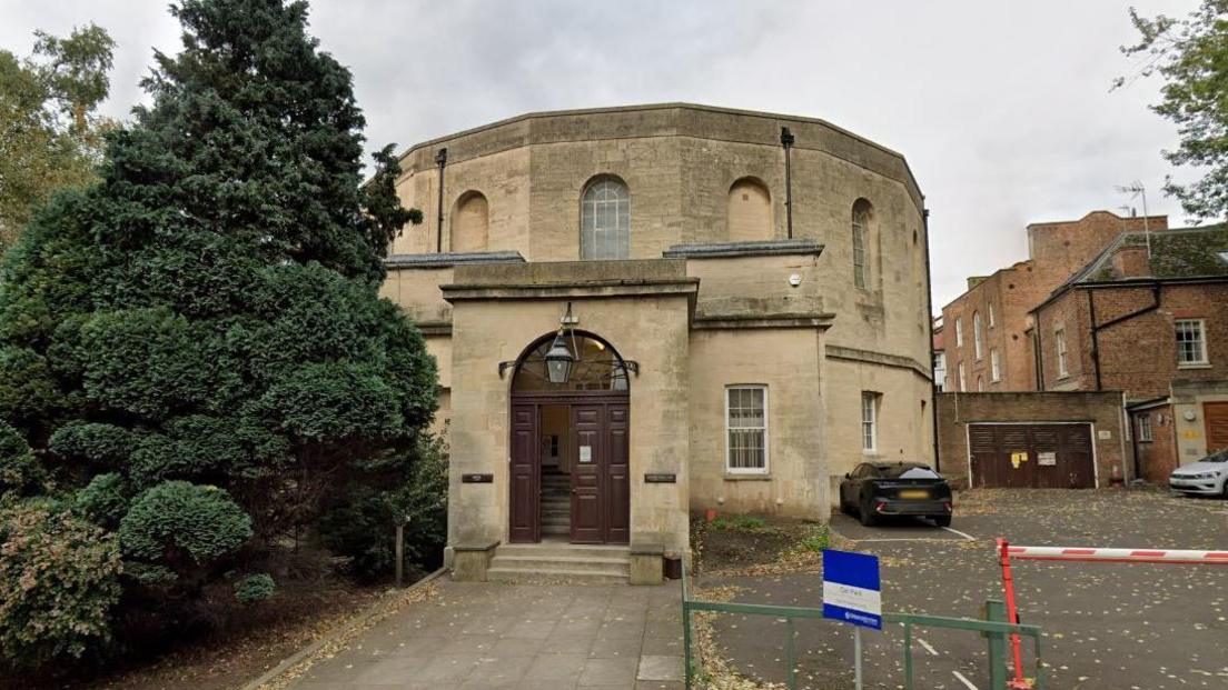 A Google street view image of Gloucester Crown Court. It is an octagonal-shaped stone building with an arched wooden door. To the left of the entrance there are large trees and to the right there is a car parking area.