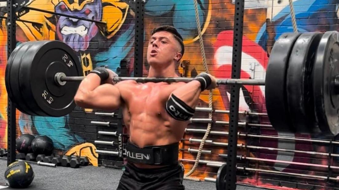 A topless man holding a weightlifting bar with several heavy weights at shoulder level