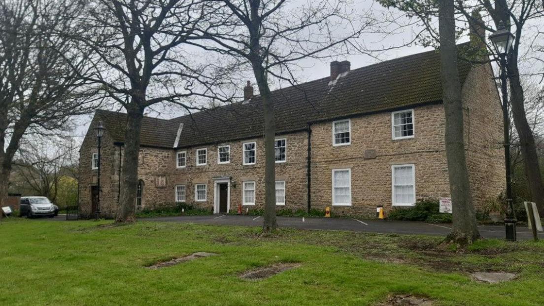 The two-storey Kepier Hall. It is a beige stone building with white windows and several trees in front of it. A car is parked on the side.