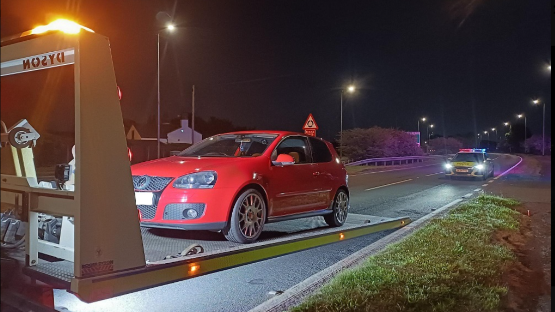 A red car is half way up a platform leading from the road onto a recovery truck. A police car with its lights flashing is a short distance behind on the same road. Grass verge is on one side and the central reservation barrier is on the other.