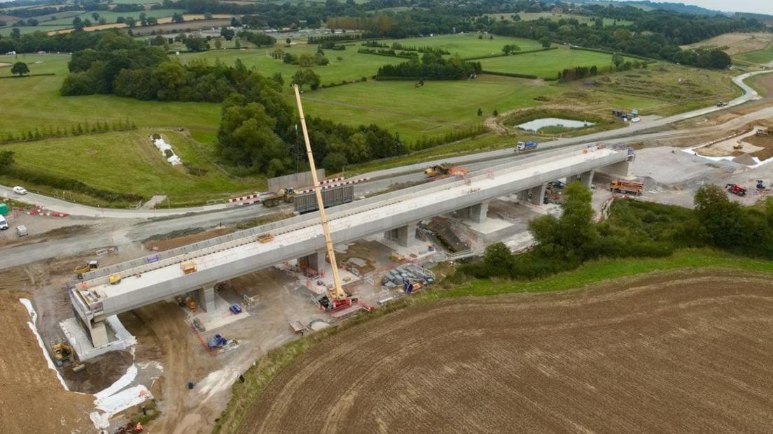 Recent shot of viaduct showing most of the scaffolding removed and grey parapets installed alongside the beams. A yellow crane is visible, with green fields and woodland in the background.