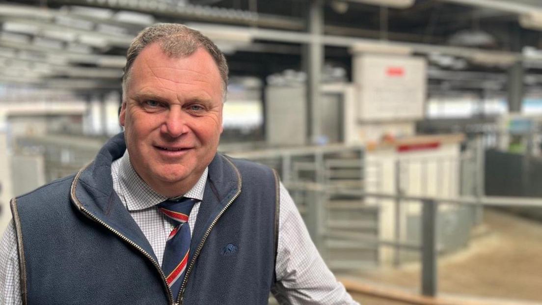 Mark Bromell in a check shirt with a striped tie and blue gilet, standing in front of the auction ring