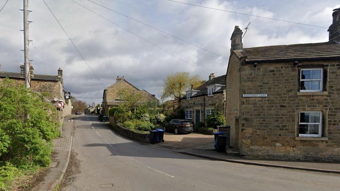 A street in a picturesque rural English village. There are stone houses, telegraph poles and a green bush.