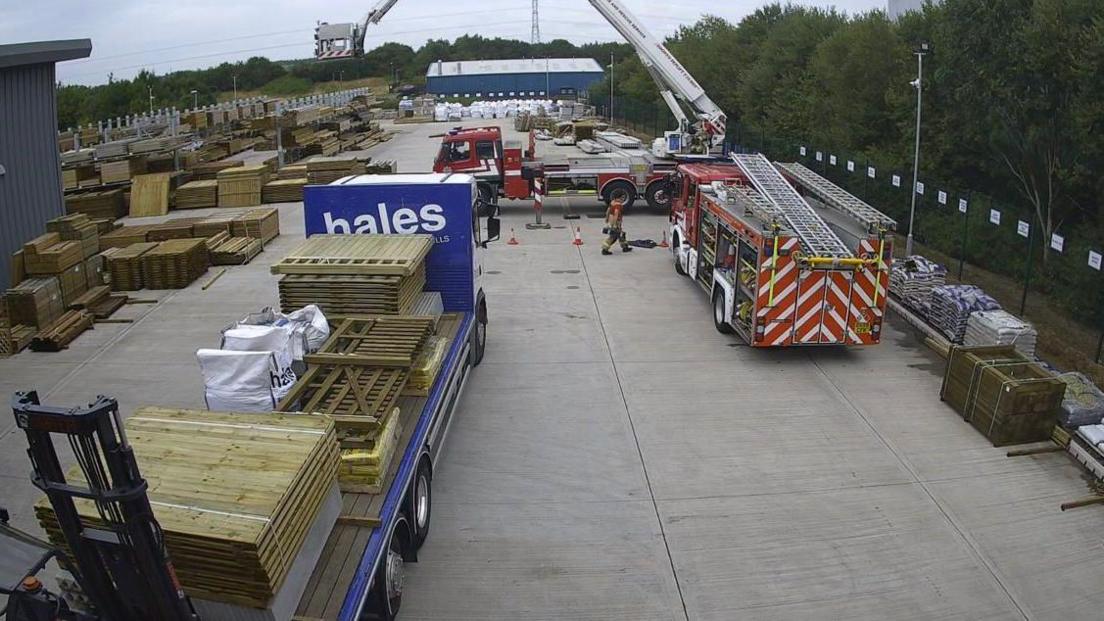 A concrete yard outside an industrial building. Piles of wooden pallets are on the trailer of a truck and on the ground on the left. Two fire engines are parked in the yard - one has its aerial appliance extended.