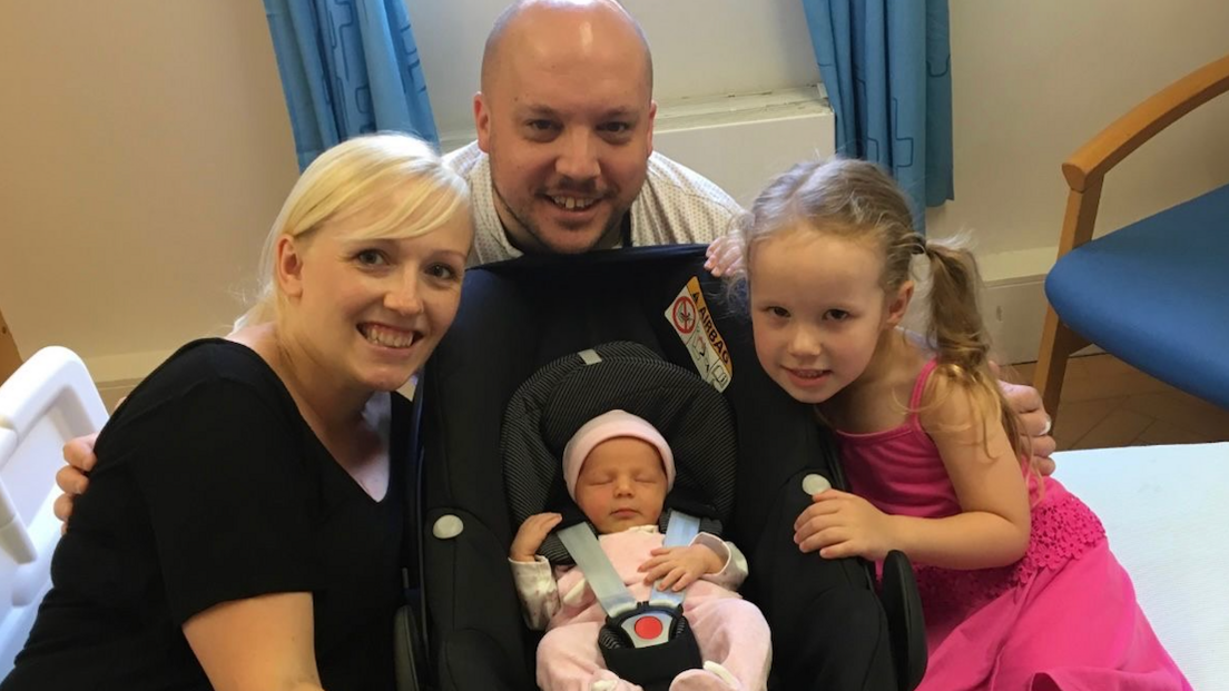 Kayleigh and husband David smile at a camera, with a baby sat in a car seat and a little girl with blonde wearing a pink dress next to them.