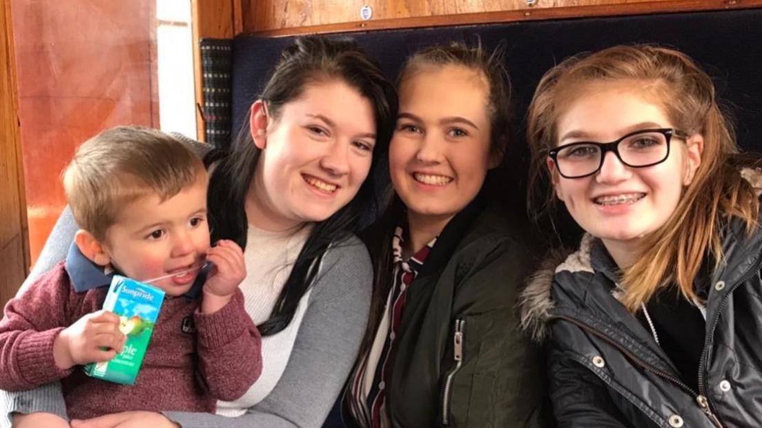 Russell family members in a train carriage. From left to right Elliot (Mrs Russell's grandson) with an apple juice pouch and burgundy jumper. Danii, the eldest daughter wearing a grey cardigan and hugging Elliot. Johdi in a green coat and on the right is Aimie with a black coat and fluffy hood.