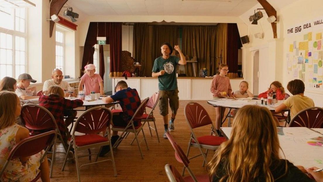 Children sitting round several round tables in a hall with a man standing at the front