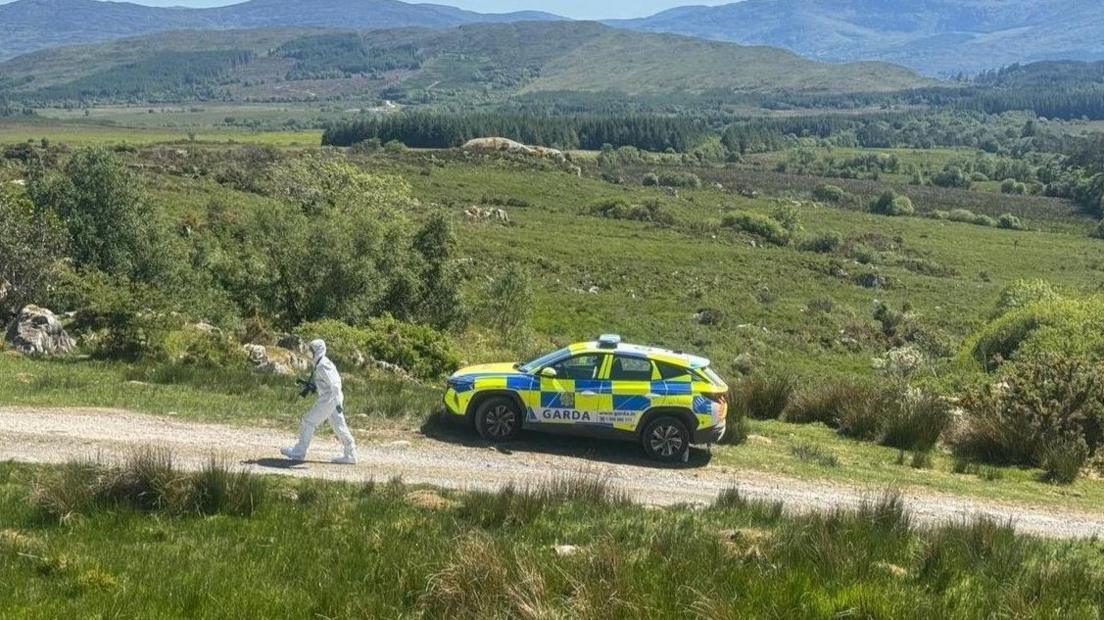 A GARDA car parked at the side of a narrow dirt road. There is a person in a white forensic suit walking up the path away from the car. Rolling green hills are in the backdrop.
