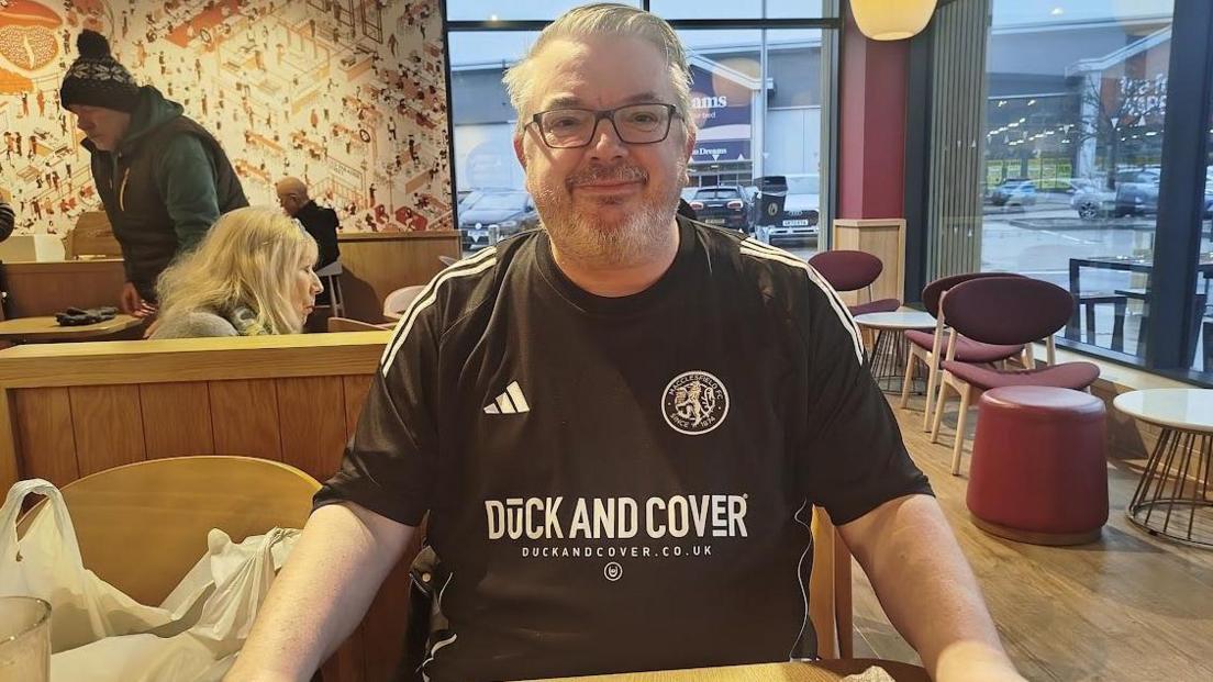 Mark Sharpley has greying hair, dark spectacles and is wearing the dark change kit of Macclesfield FC withshirt sponsor Duck and Cover emblazoned on it. He is sitting in a branch of Costa Coffee.