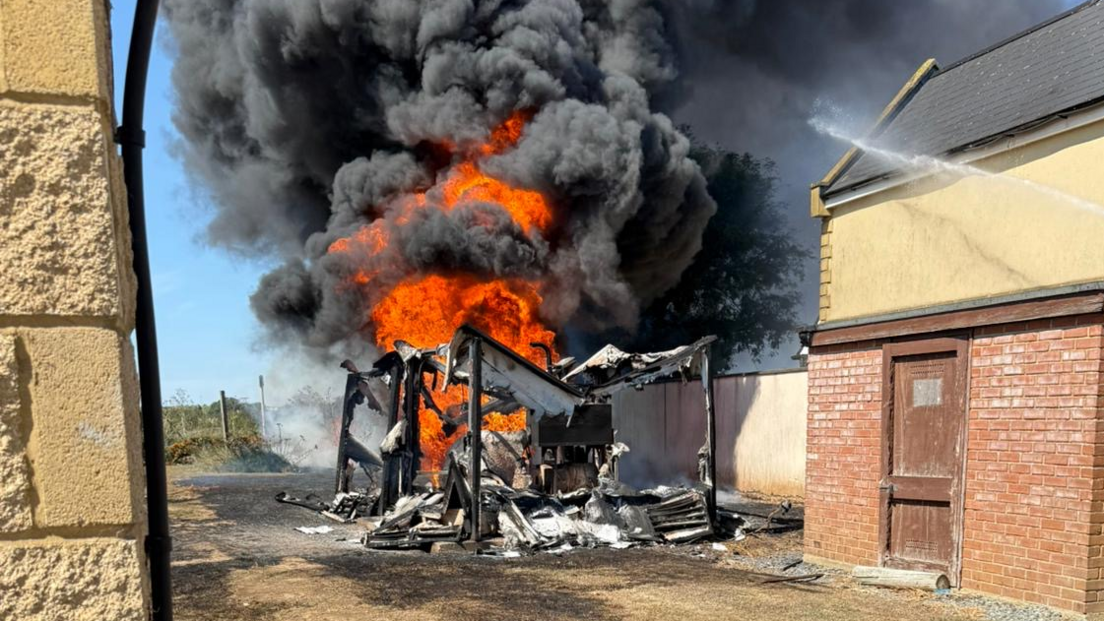 Water from a hose is blasted onto a large fire, which has destroyed a small building. Thick black smoke is billowing out from the blaze. There are two other buildings next to where the fire is.