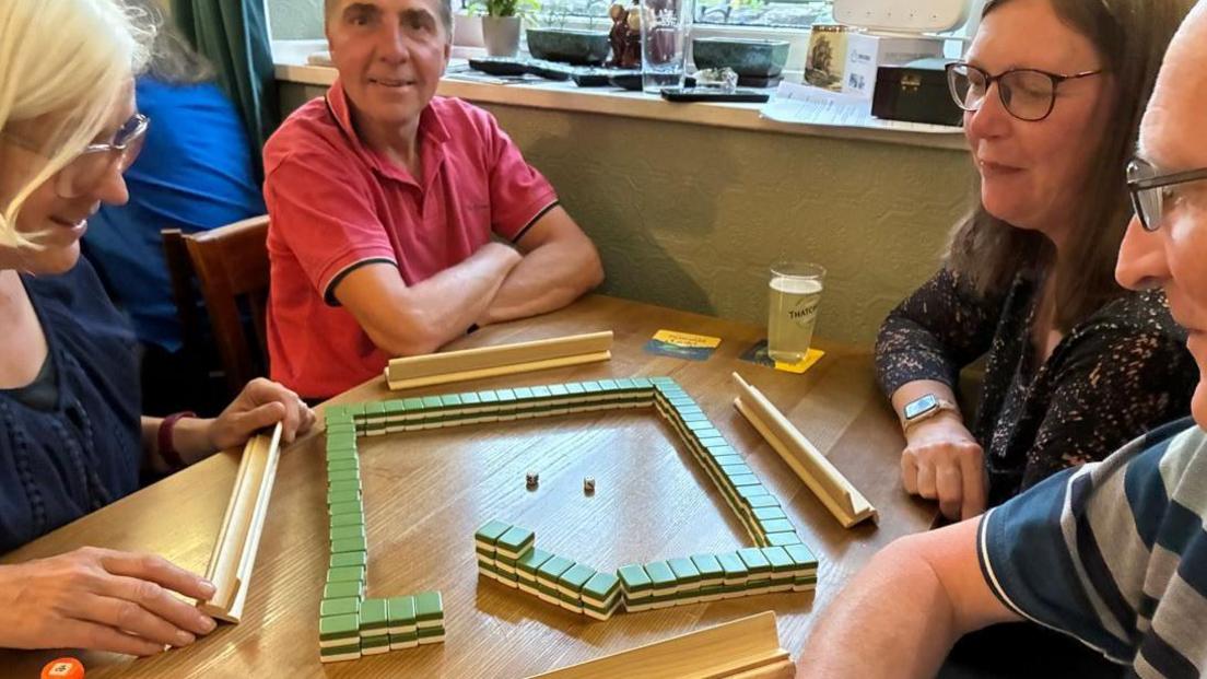 A group of people enjoying Mahjong. There are two women and two men sitting at a table with the game set out in front of them. One of the men is smiling directly at the camera.