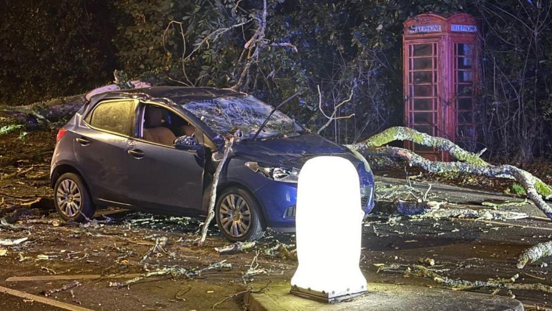 Car with tree branches on it and  smashed windscreen in a road with a phone box.