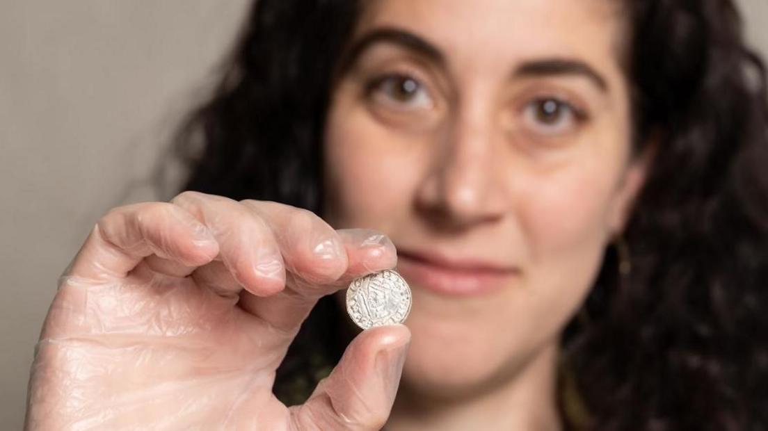 Amal Khreisheh, who is wearing a clear latex glove, holds up one of the coins between their thumb and forefinger. Their face is out of focus in the background.