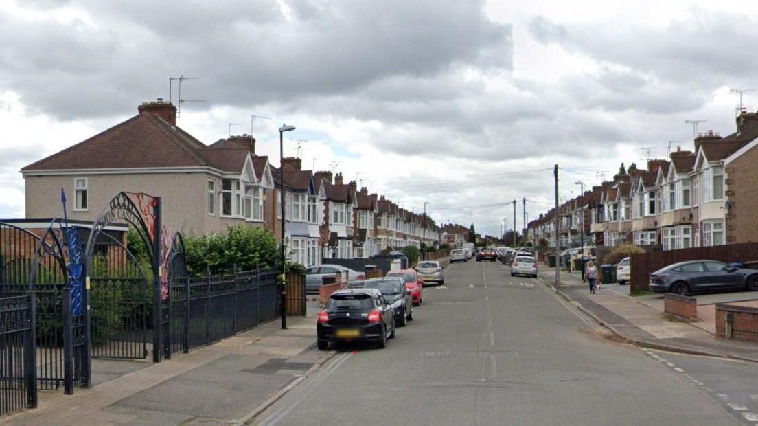 A Street with a black metal archway to the left which also has red and blue features, on the road there are a number of parked cars and terraced housing either side