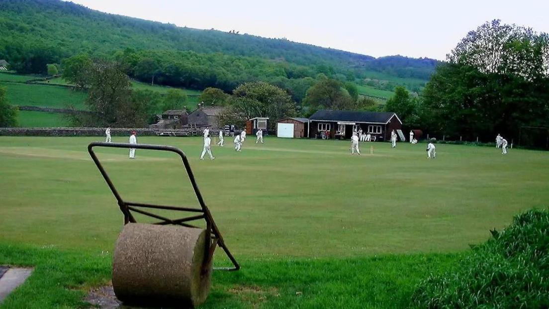 A cricket field with about 14 players dressed in white. In the background are a cricket club house, a shed, a farmhouse and green hills.  In the front of the picture is a lawn roller. 