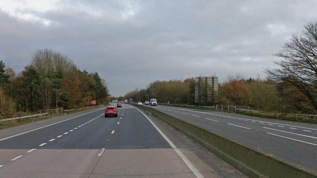 A section of the M54 motorway. Cars and lorries can be seen in the background. The road is grey with white dotted lines and trees either side.