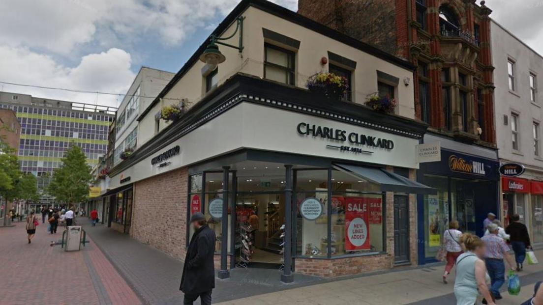 The Charles Clinkard store is a two-storey corner building. It has a black and white sign. People are walking by the shop.