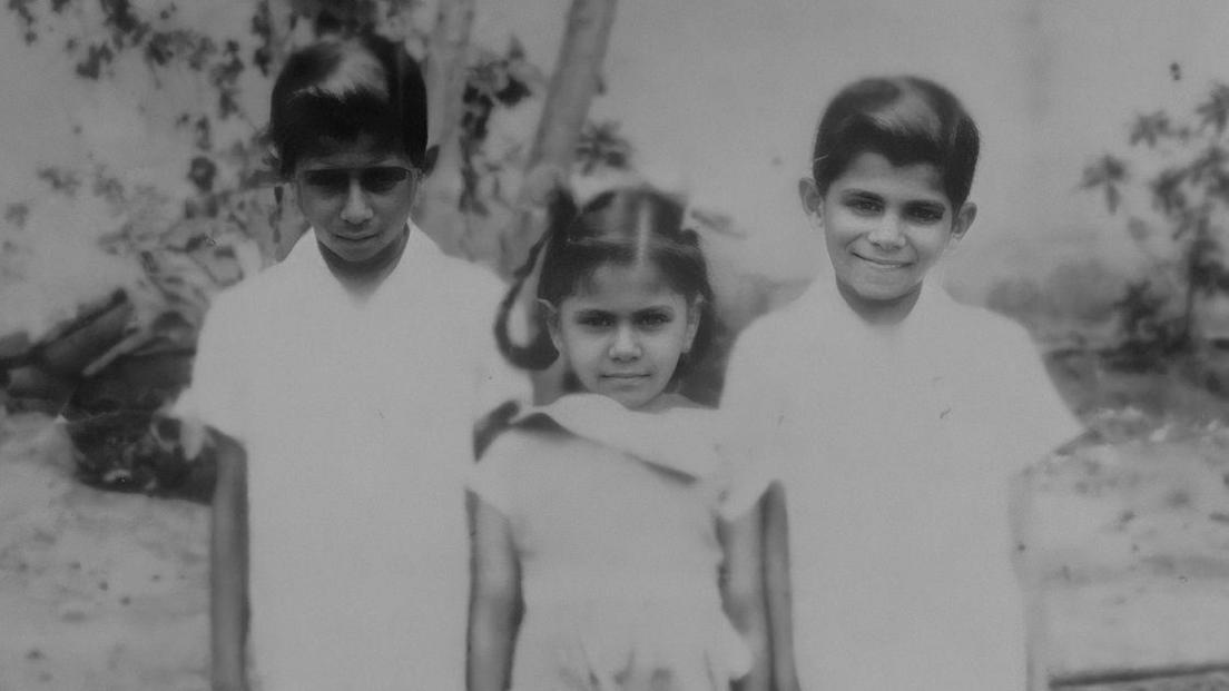 A black and white photo showing a young Tara Mistry standing in the middle of two boys. The boys are wearing white shirts while Tara wears a white dress.