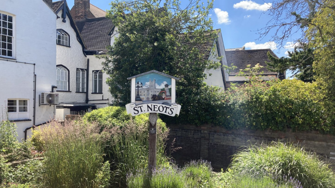 A decorative town sign reading "St Neots" stands in front of some shrubs and trees and historic white houses with tiled roofs. The sky is blue with fluffy white clouds. 