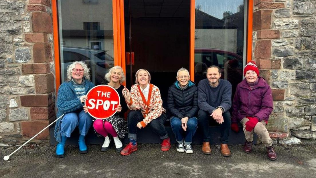 Team Spot smiling at the camera, as they sit outside The Old Mart in Victoria Road. From left to right: Zoe Mander, Ceri Hutton, holding a red sign reading The Spot, Jennie Dennett, who holds the keys, Helen Shacklady, Simon Wand and Stephanie Sexton. 