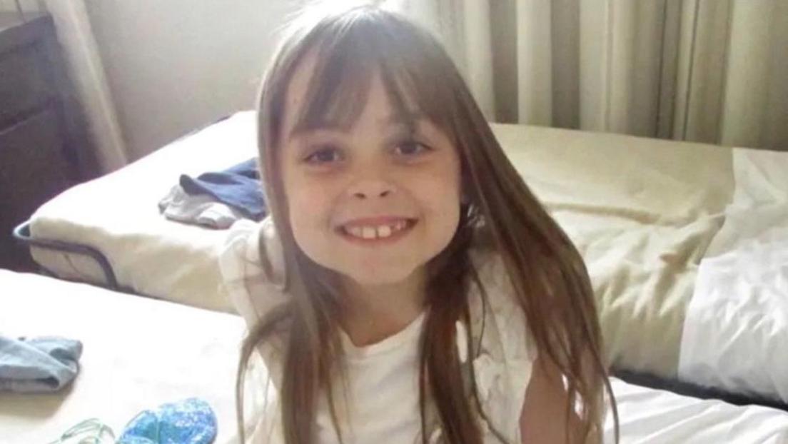 Saffie Roussos is sitting on a bed and smiling directly at the camera.