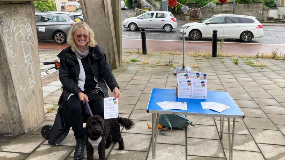 A woman sits on a chair with a black dog on a lead next to a table with leaflets. She is sitting beneath a flyover on a main road. 