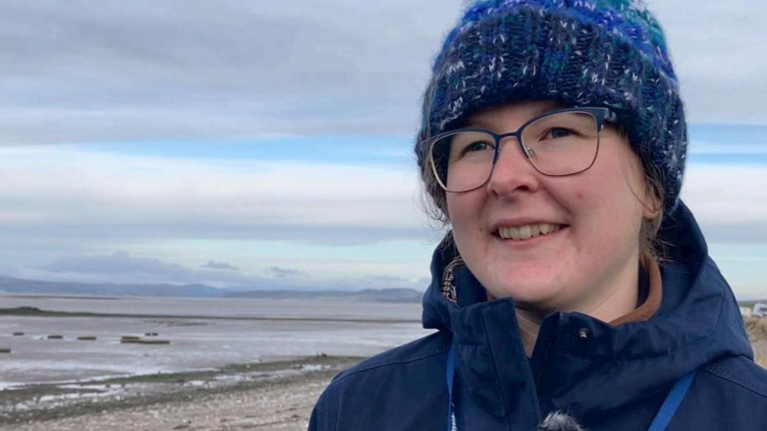 Eleanor Brown, wearing a blue bobble hat, glasses and a dark blue coat, stands in front of Lancashire's salt marshes.