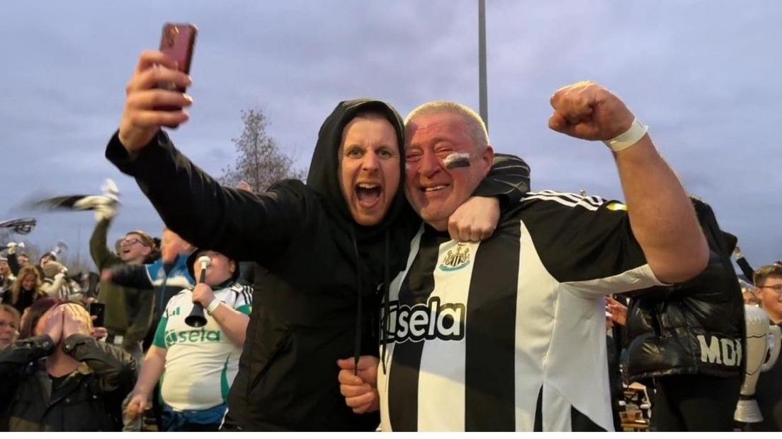 A scene from the Newcastle fan zone. On the left a young man in a black hoodie holds aloft a mobile phone. His left arm is around the shoulder of an older man wearing a Newcastle United strip and who appear to be crying. 