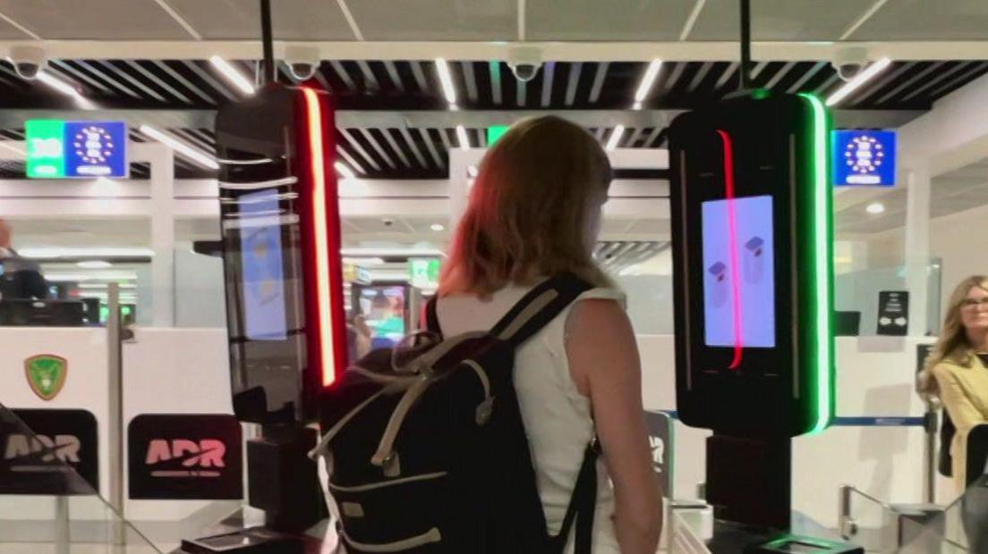 A woman with long light brown hair and a rucksack standing at an electronic border control gate, which is recording a scan of her face.