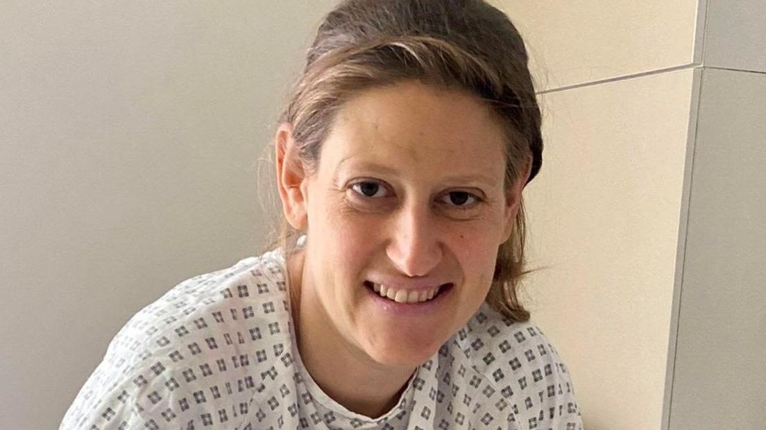 A woman looks at the camera. She has dark blonde hair, pulled behind her head and wears a hospital gown of white with a dark diamond pattern on it. 