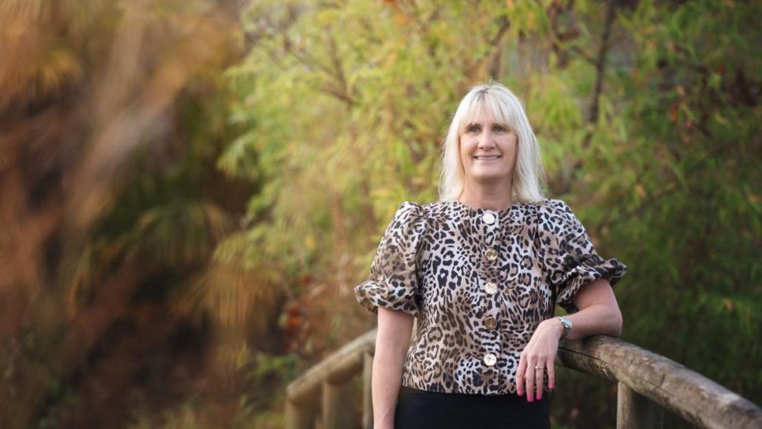 Rebecca Brewer seen wearing an animal print top as she leans on a wooden fence with trees surrounding her.